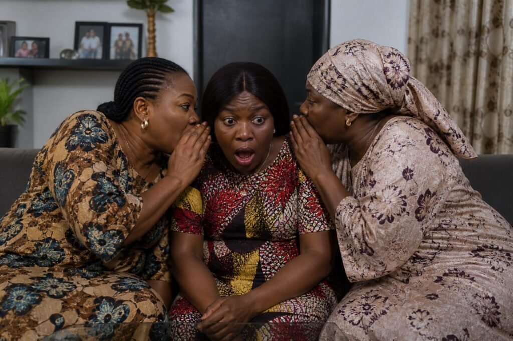 Three Nigerian women sitting close together whispering and reacting with surprise in a living room.