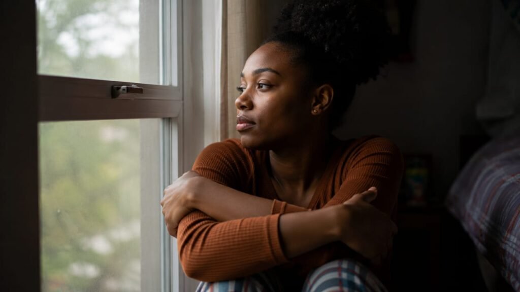 A young woman sits by a window in soft daylight, holding herself closely while staring outside in deep thought.